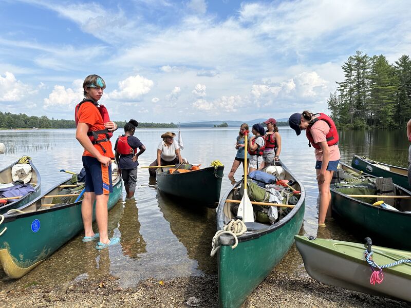 The image shows a group of people with canoes on a lake shore. Some are standing near the canoes, while others are in the water. The canoes are of different colors, and the people are wearing life jackets. The background includes trees and a cloudy sky. It seems like they are preparing for a canoe trip or have just finished one.